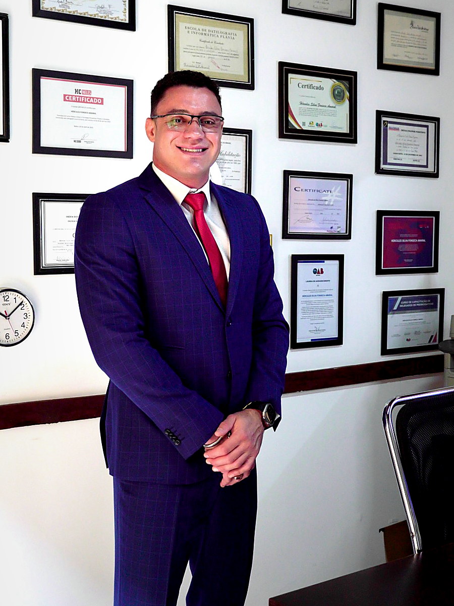 Hércules S. Fonseca Amaral de pé em seu escritório, diante da parede de diplomas e certificações
